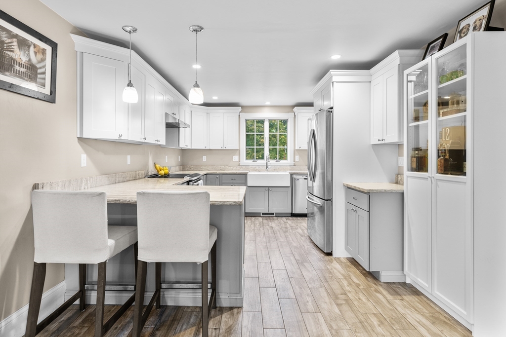 Bright white kitchen with a large island, two stools, stainless appliances, and a farmhouse sink beneath the window.