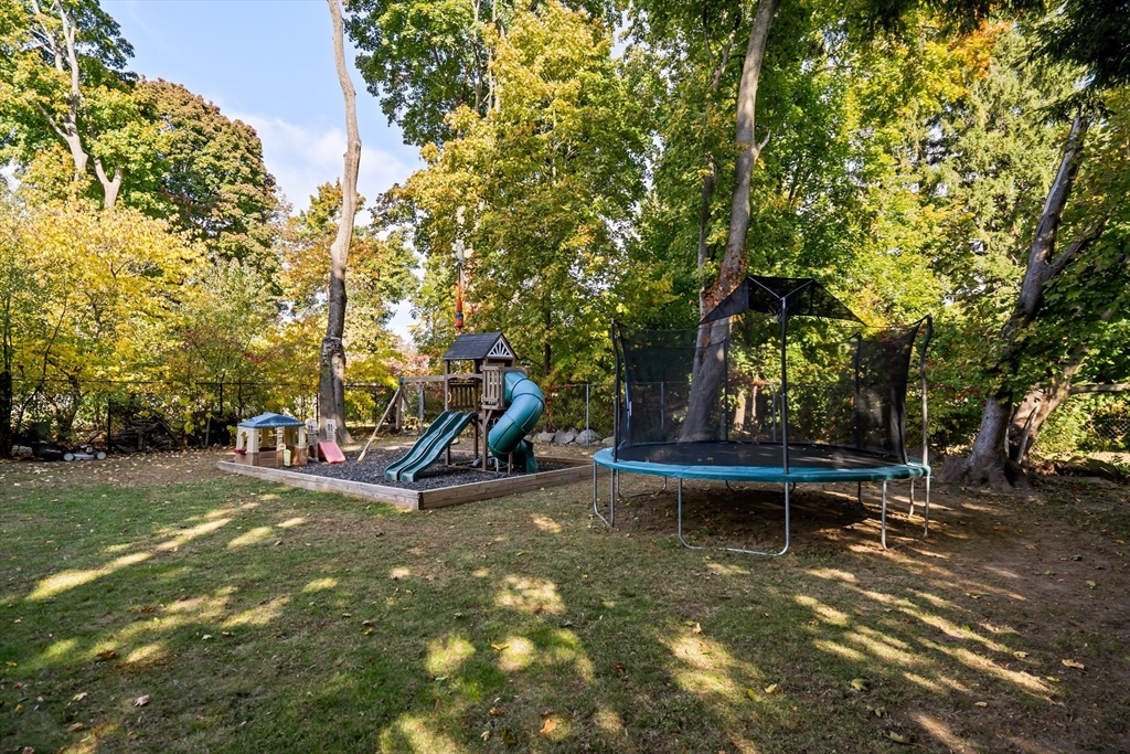 Backyard with a children's playset, slides, and an enclosed trampoline beneath mature trees.