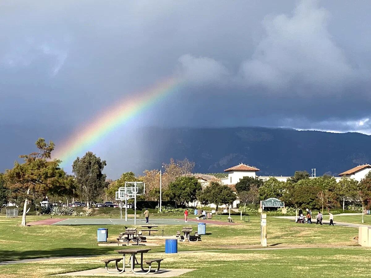 Rainbow over a neighborhood park with basketball courts, picnic tables, and mountains.