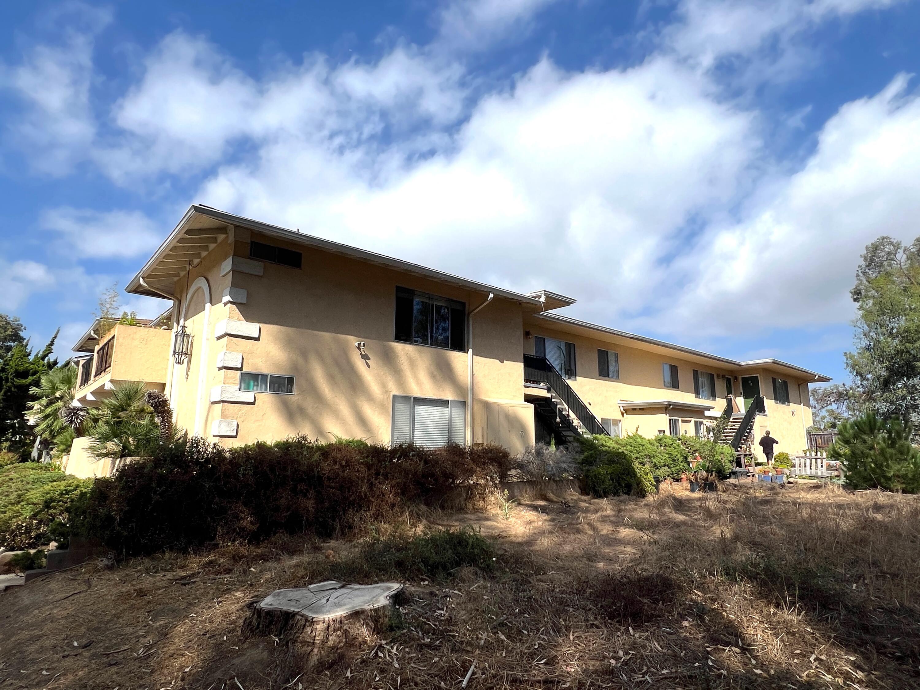 Two-story tan stucco building with exterior staircases above a dry, overgrown slope.