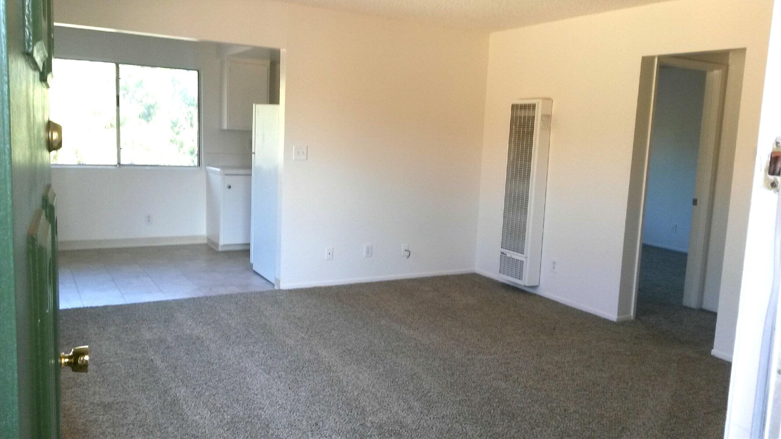 Empty living room with gray carpet, wall heater, and tiled kitchen area.
