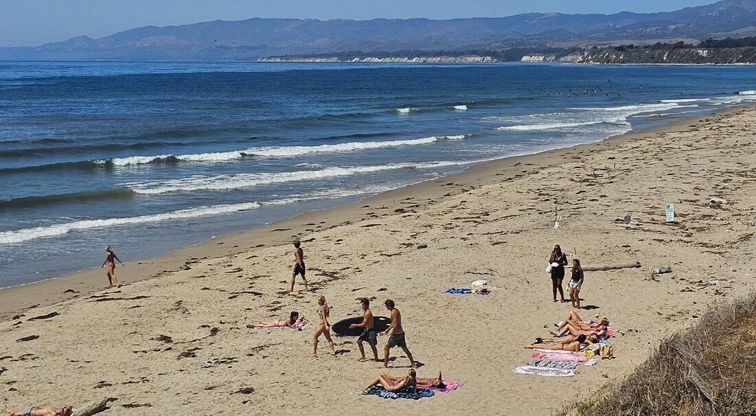 Sandy beach with beachgoers, gentle waves, and distant mountains across the water.