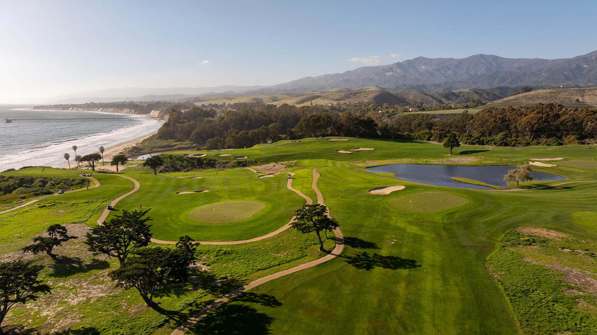 Coastal golf course with ocean shoreline, pond, and mountains in the distance.
