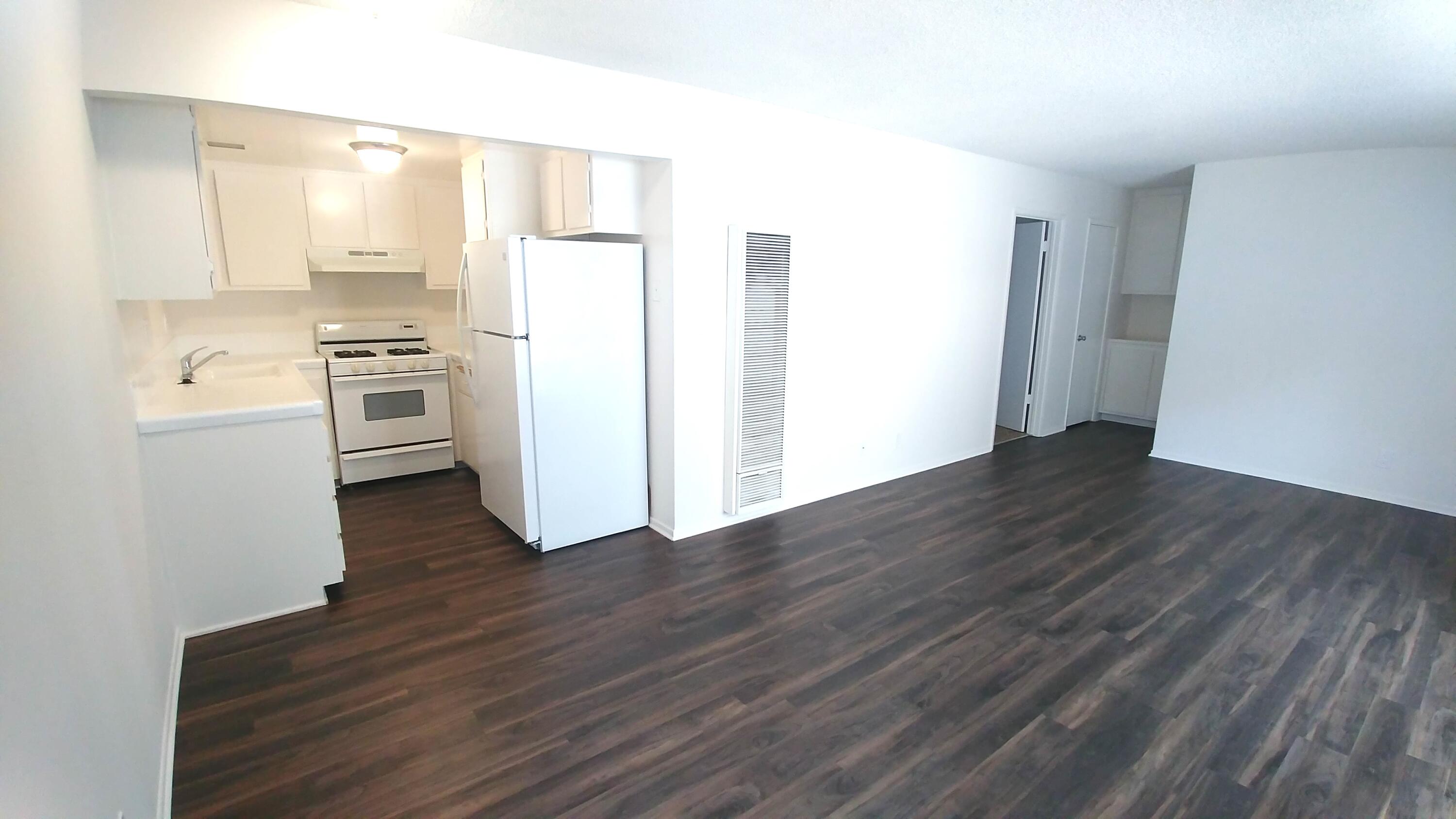 Empty apartment living area with a compact white kitchen and dark wood-look flooring.