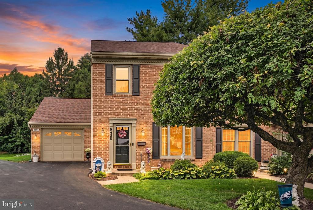 Two-story brick house with an attached garage, dark shutters, and a large leafy tree at dusk.
