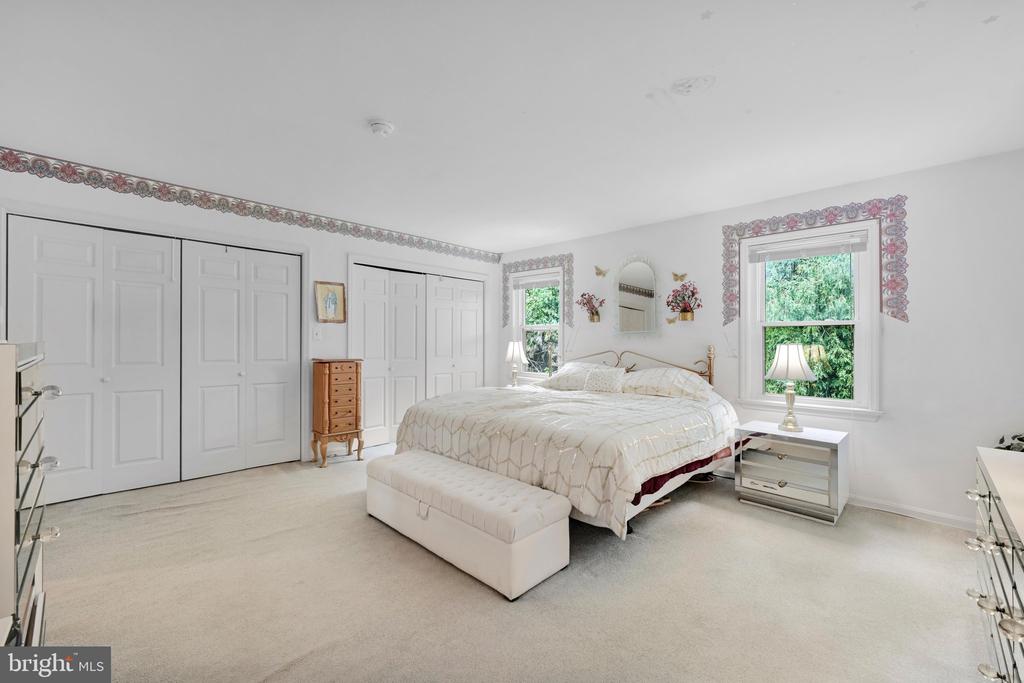 Bright bedroom with a large bed, upholstered bench, white closet doors, and windows overlooking greenery.