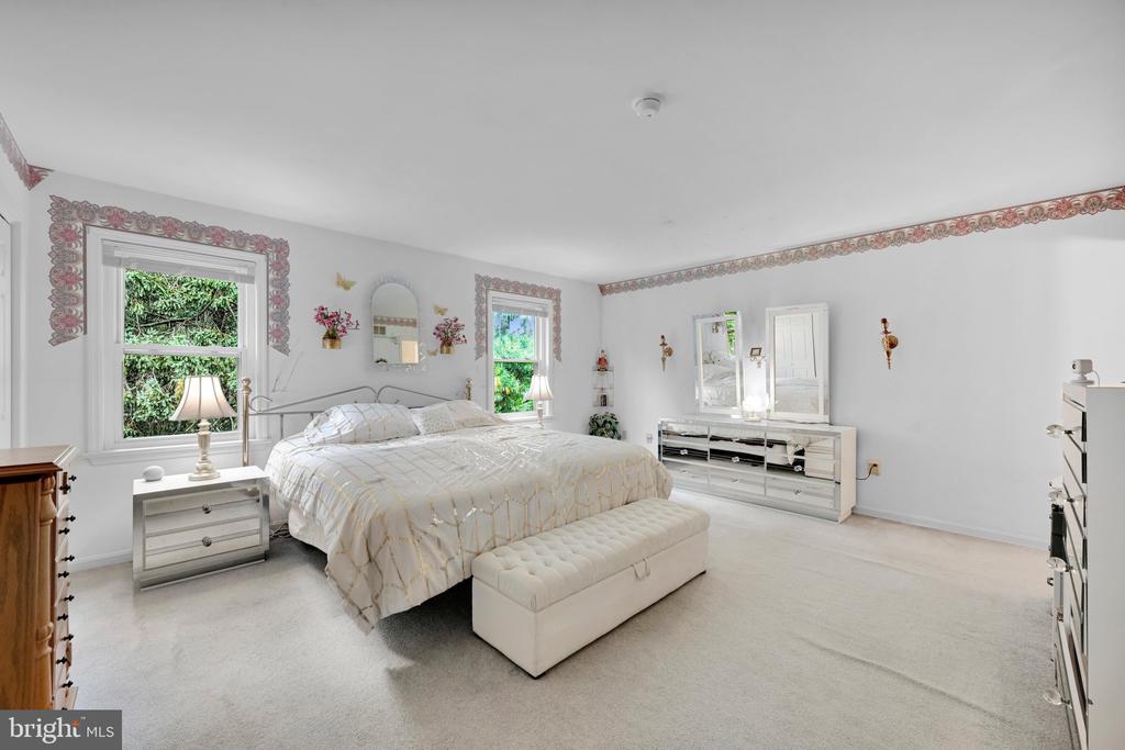 Bedroom with a large bed, upholstered bench, mirrored dresser, and two windows overlooking greenery.