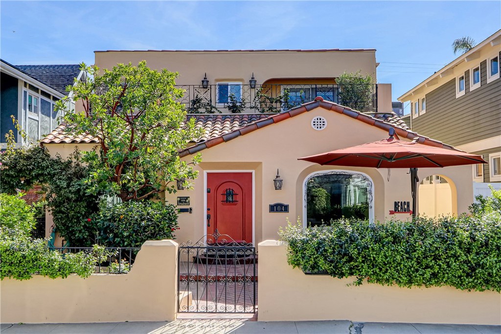 Beige stucco home with a red front door, tiled roof, and gated, hedge-lined entrance.