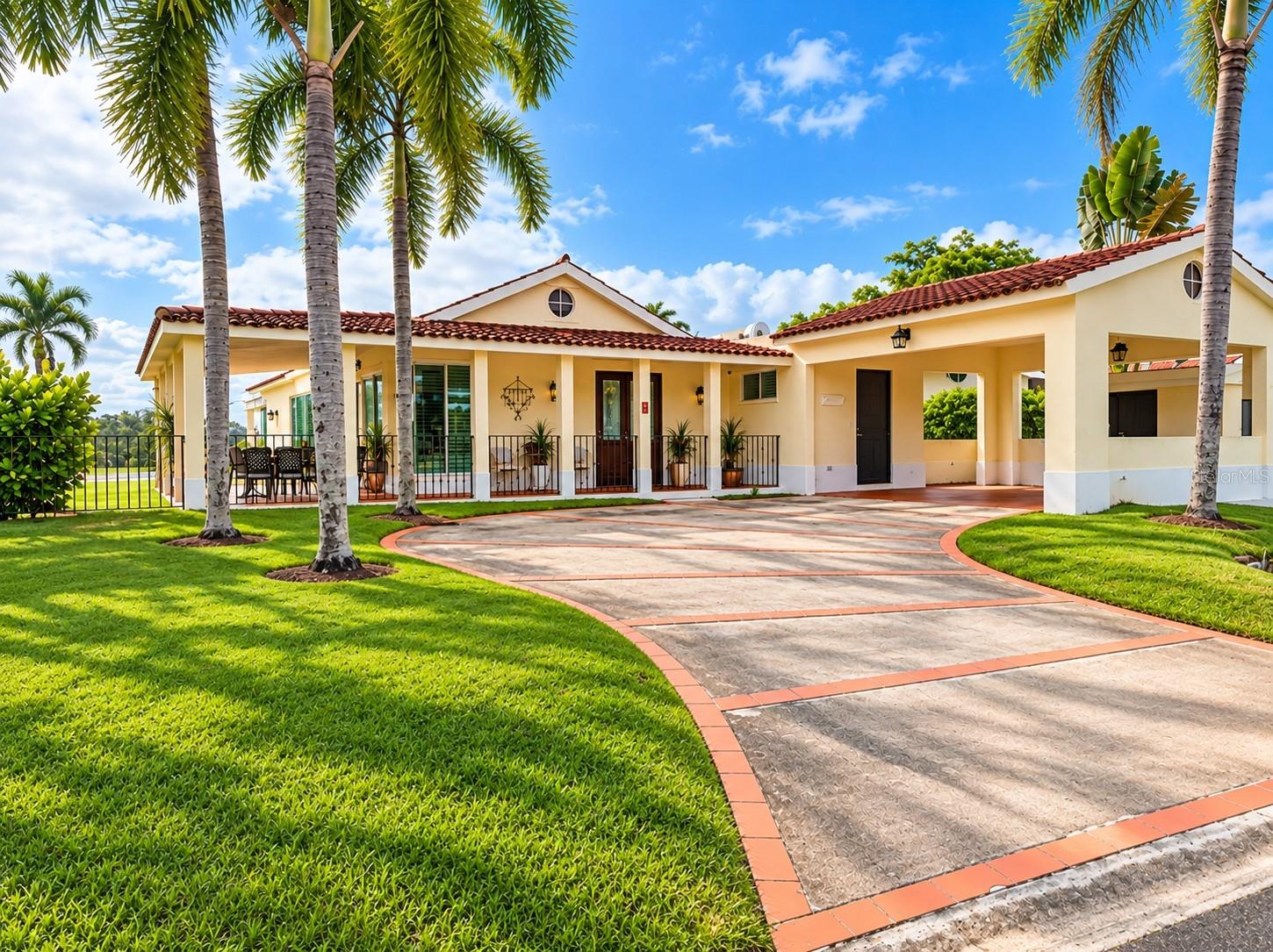 Cream-colored home with red tile roofs, a broad curved driveway, green lawn, and tall palm trees.