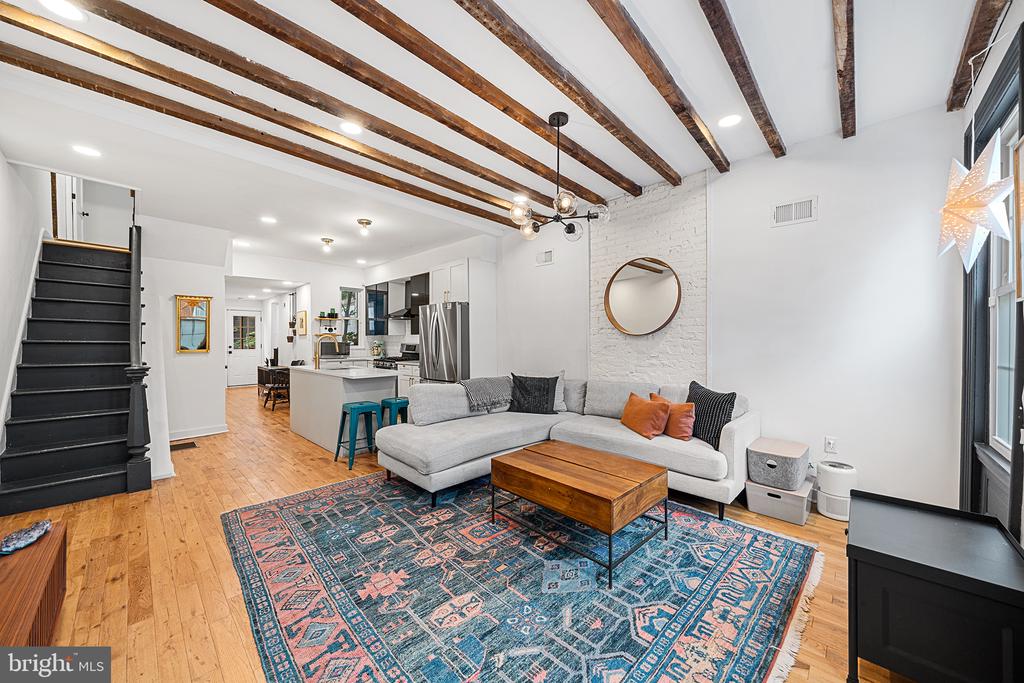 Open-plan living room with exposed ceiling beams, a white brick wall, and a gray sectional.