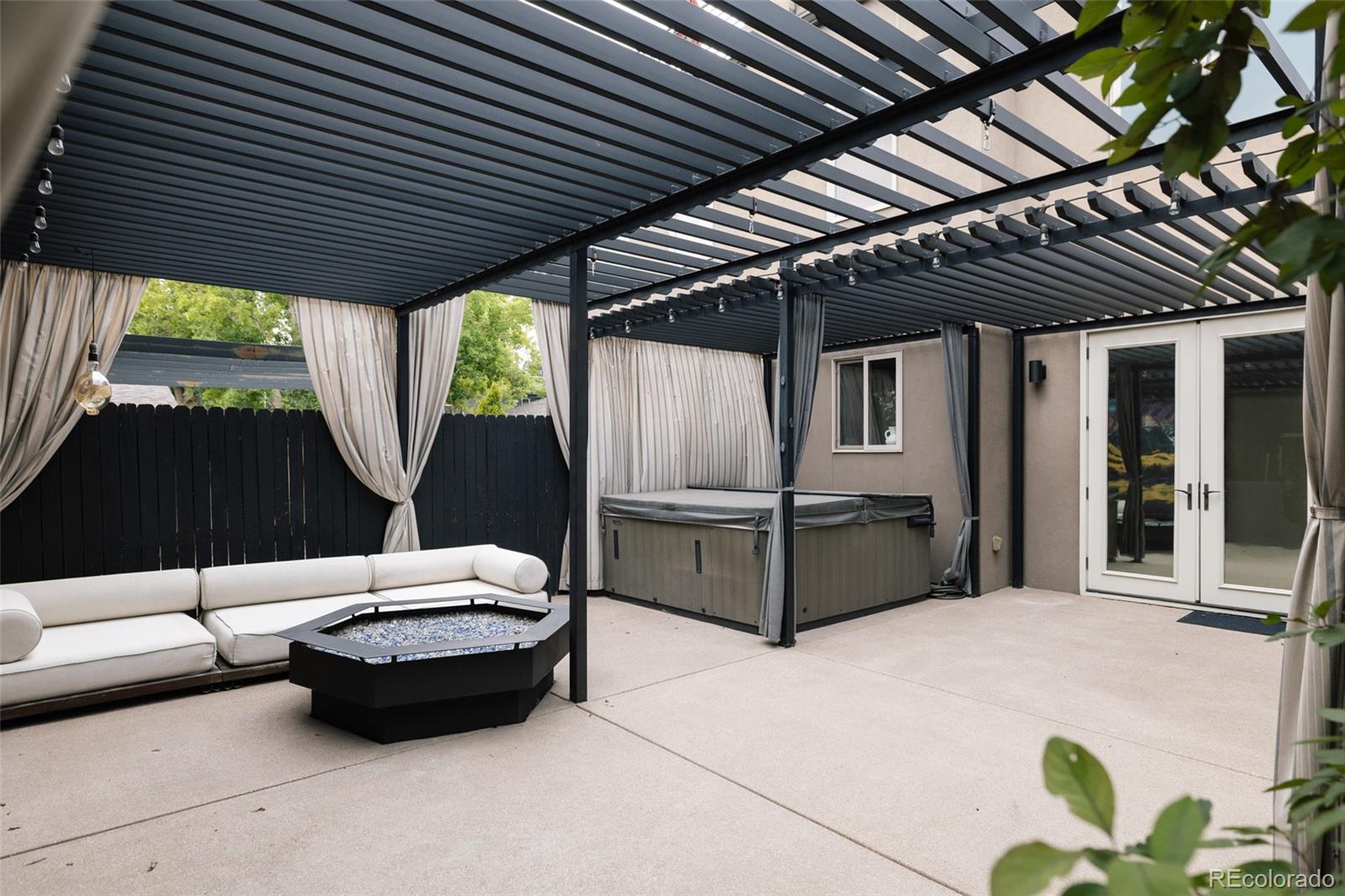 Covered patio with a hot tub, white sectional seating, and fire pit beneath a pergola.