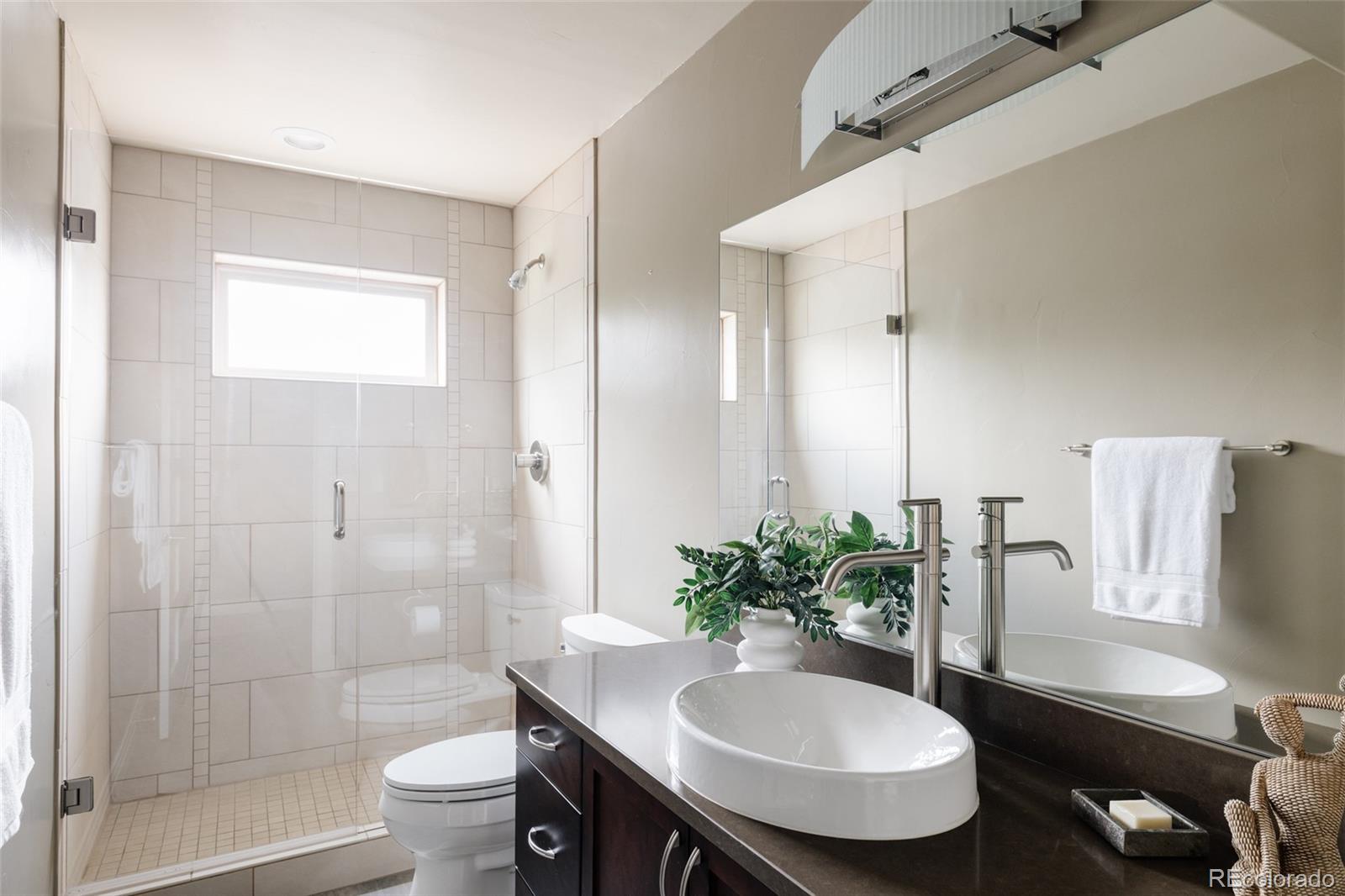 Bathroom with a tiled glass shower, high window, and dark vanity with a white vessel sink.