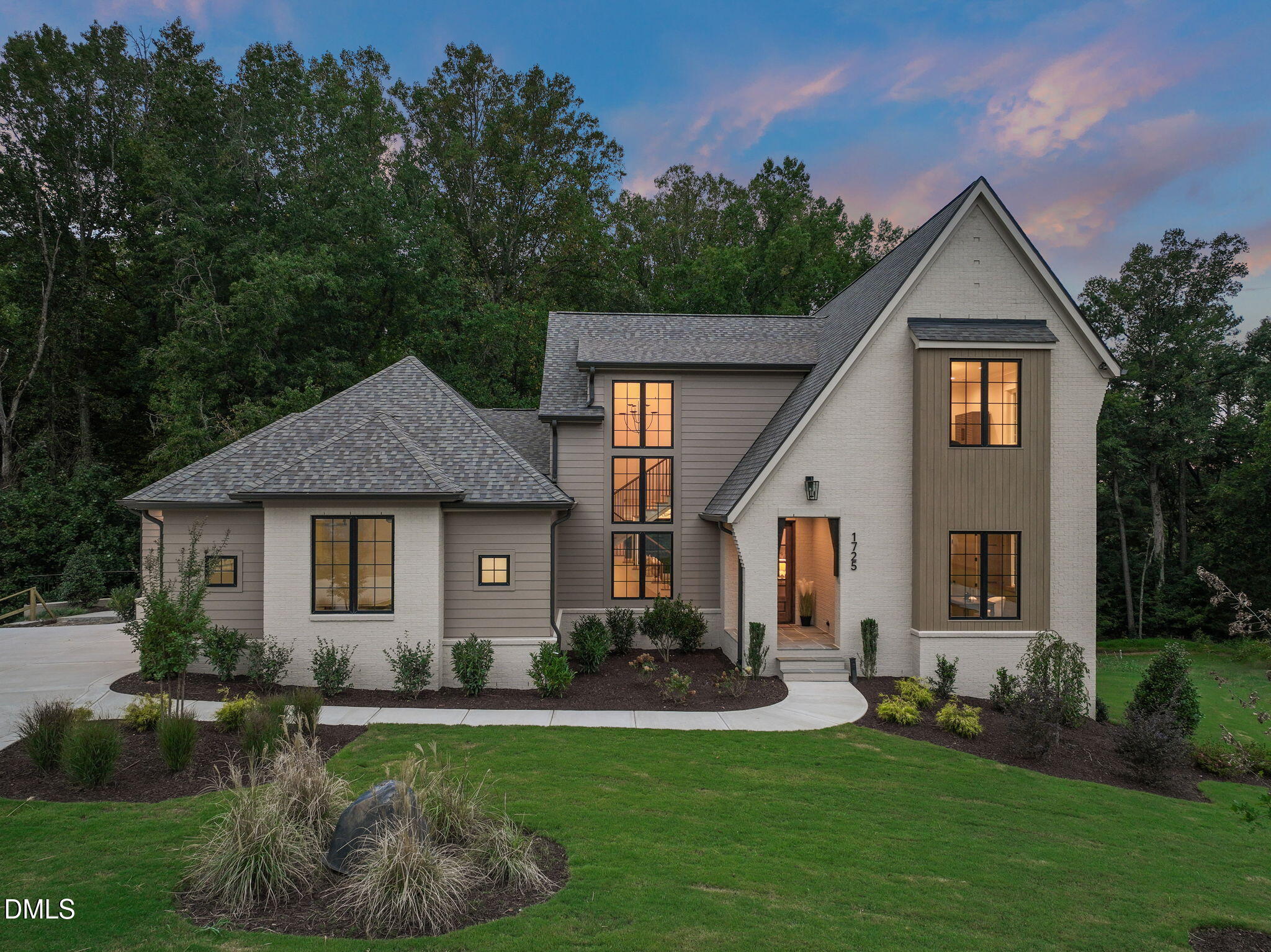 Modern gabled home with illuminated windows, landscaped lawn, and wooded backdrop at dusk.