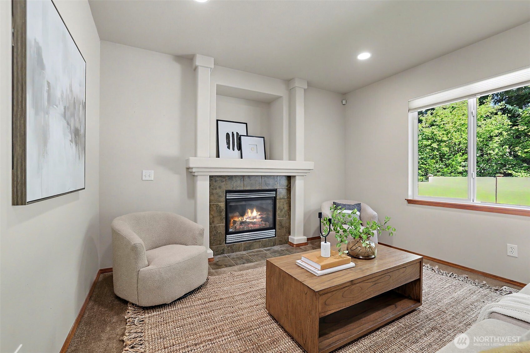 Staged living room with a tiled fireplace, neutral seating, and a large window overlooking greenery.