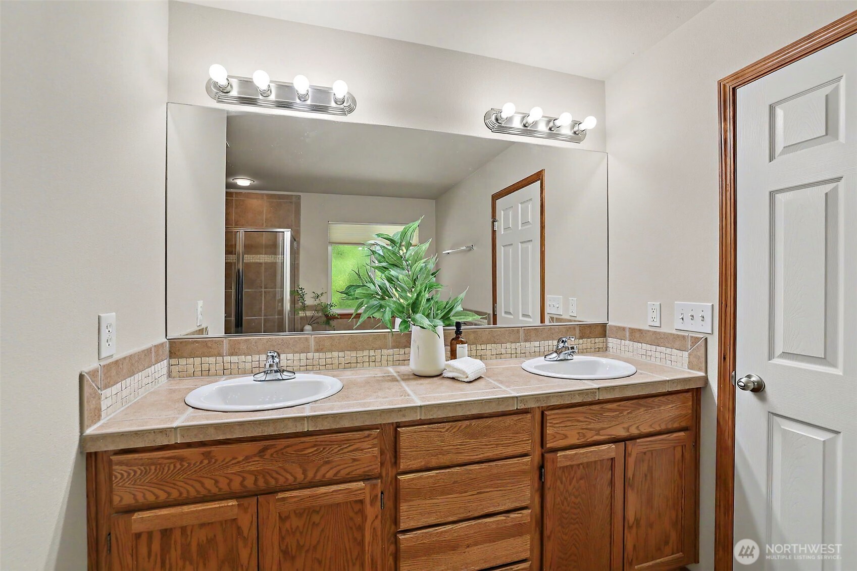 Bathroom vanity with two sinks, oak cabinets, and a large mirror reflecting a shower and window.