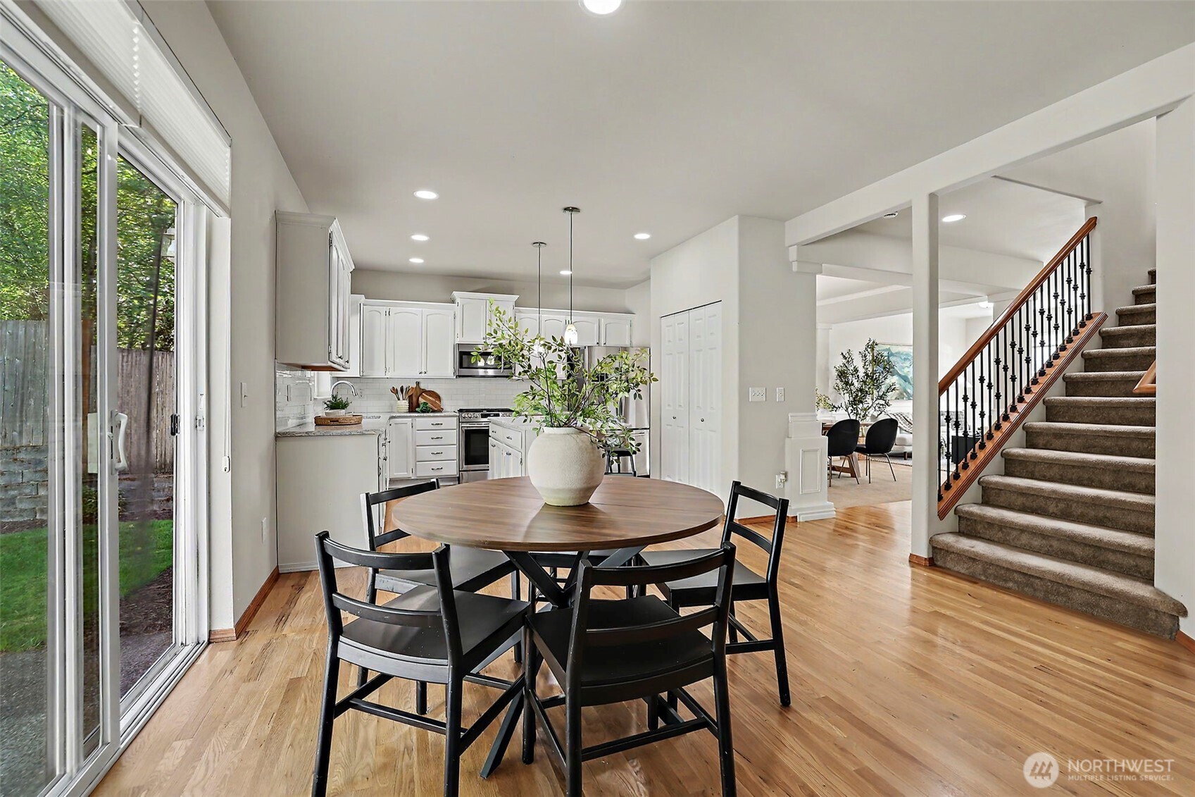 Dining area with round table beside a white kitchen, sliding glass doors, and carpeted staircase.