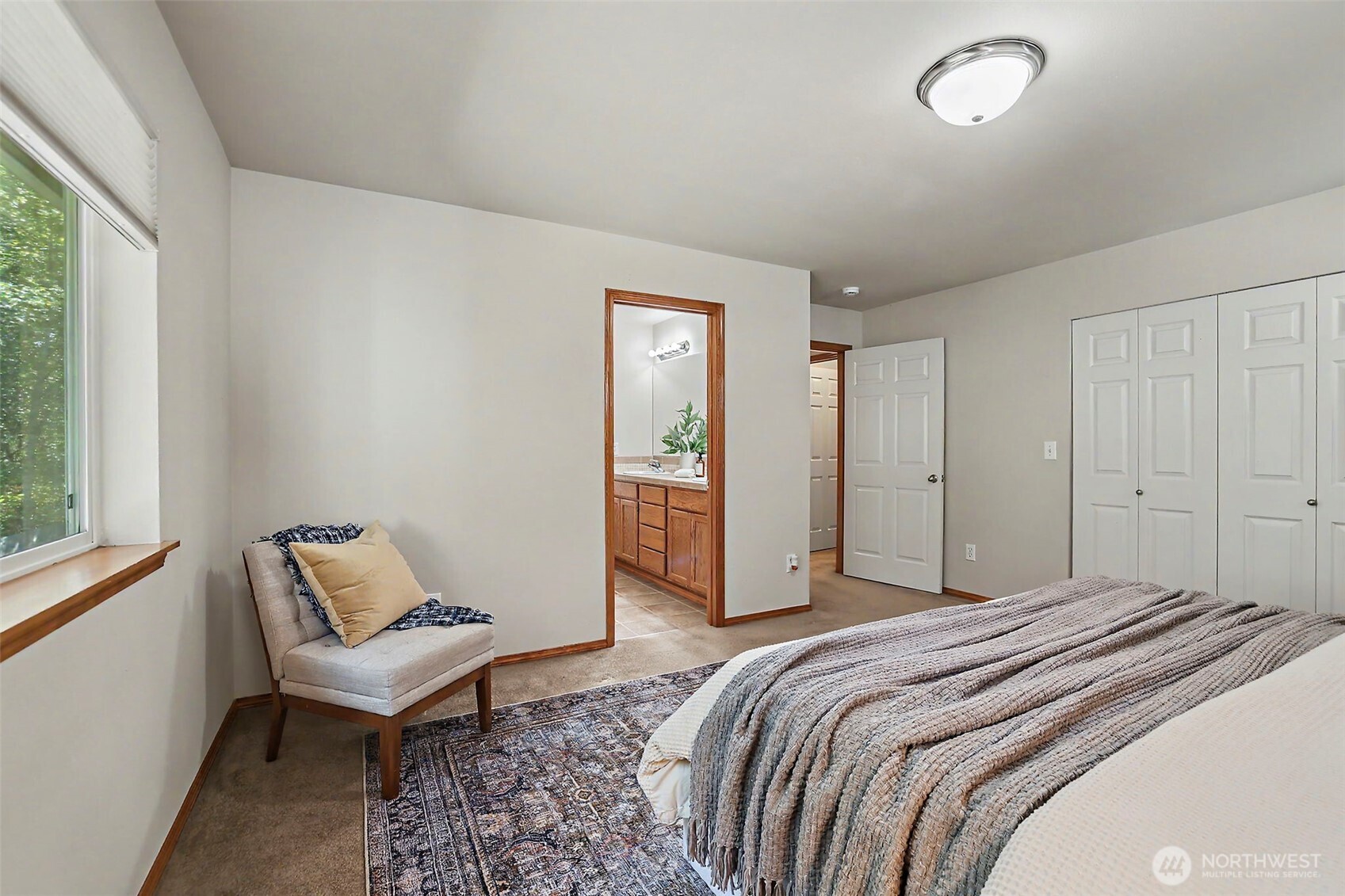 Bedroom with a large window, cushioned chair, patterned rug, and doorway to an en-suite bathroom.