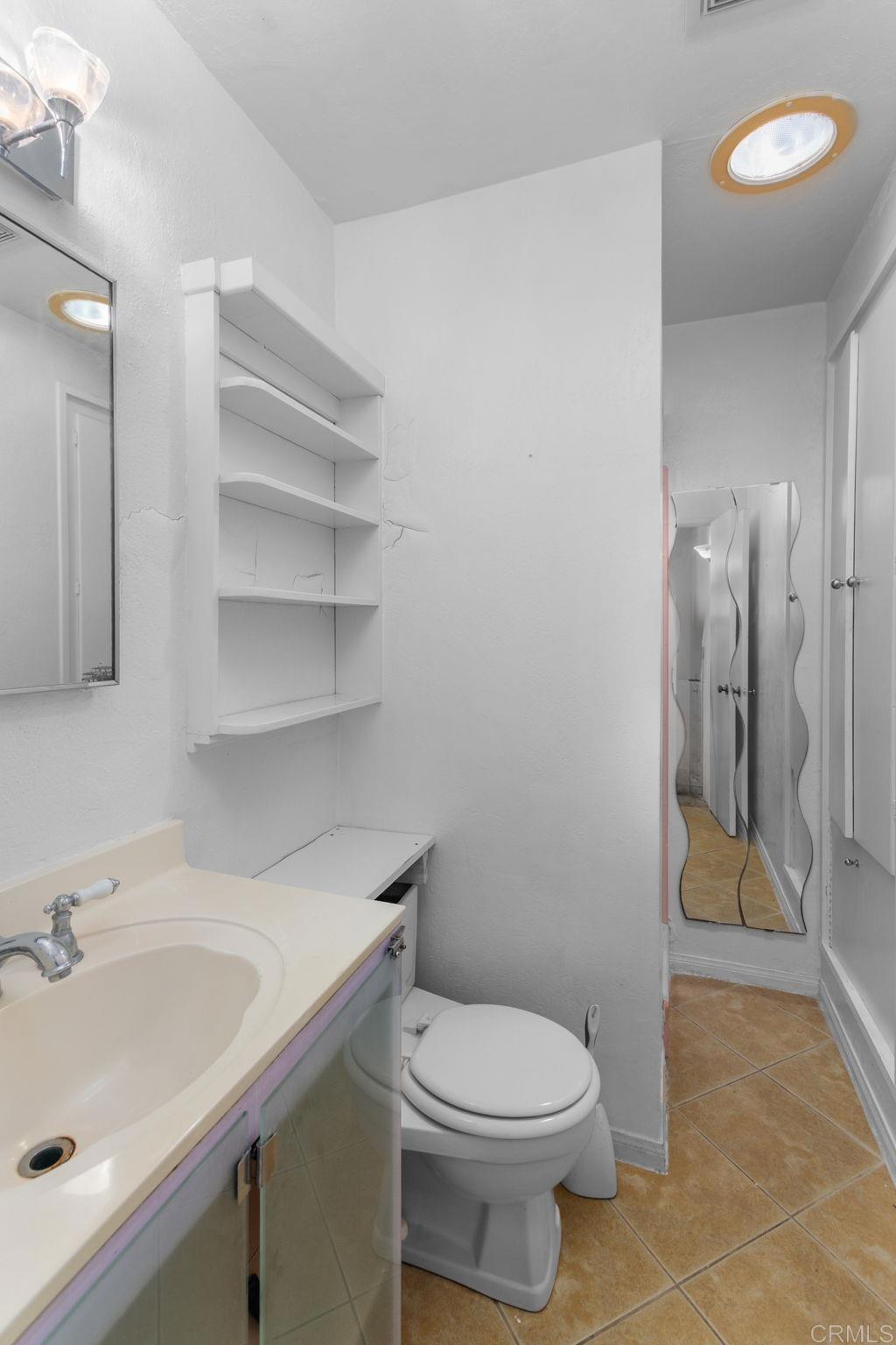 Vacant bathroom with a cream vanity, toilet, built-in shelves, and tan tile floor.