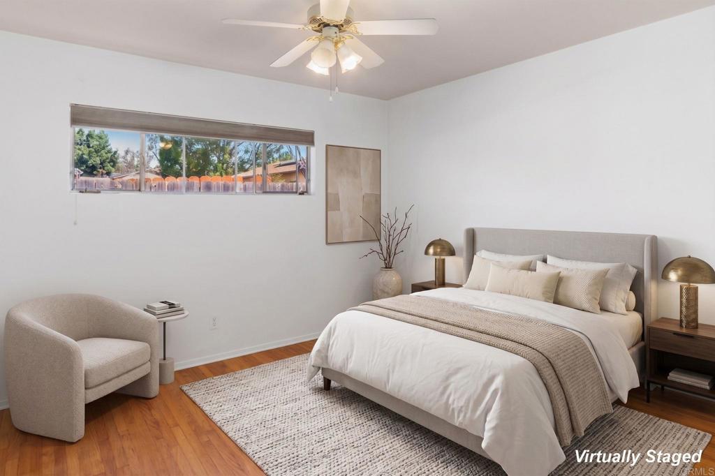 Virtually staged bedroom with a neutral bed, hardwood floors, and a wide window overlooking trees.