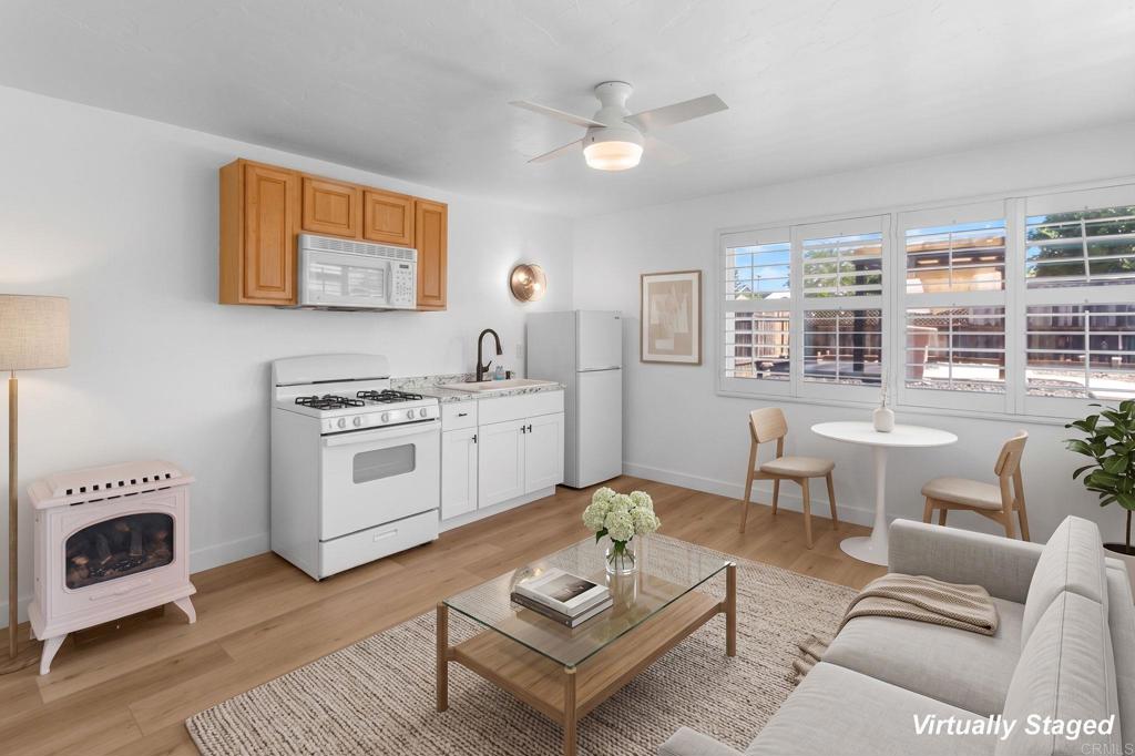 Staged living room and kitchenette with white appliances, wood cabinets, and wide shuttered windows.