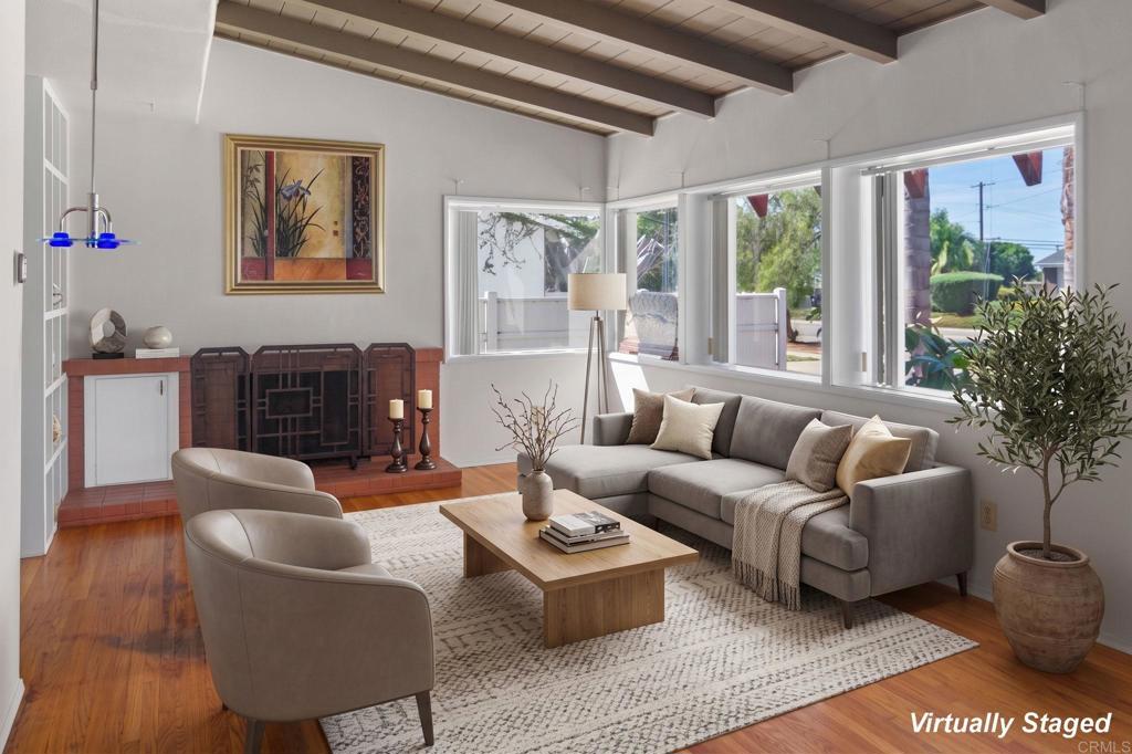 Living room with exposed ceiling beams, a brick fireplace, and large windows overlooking greenery.