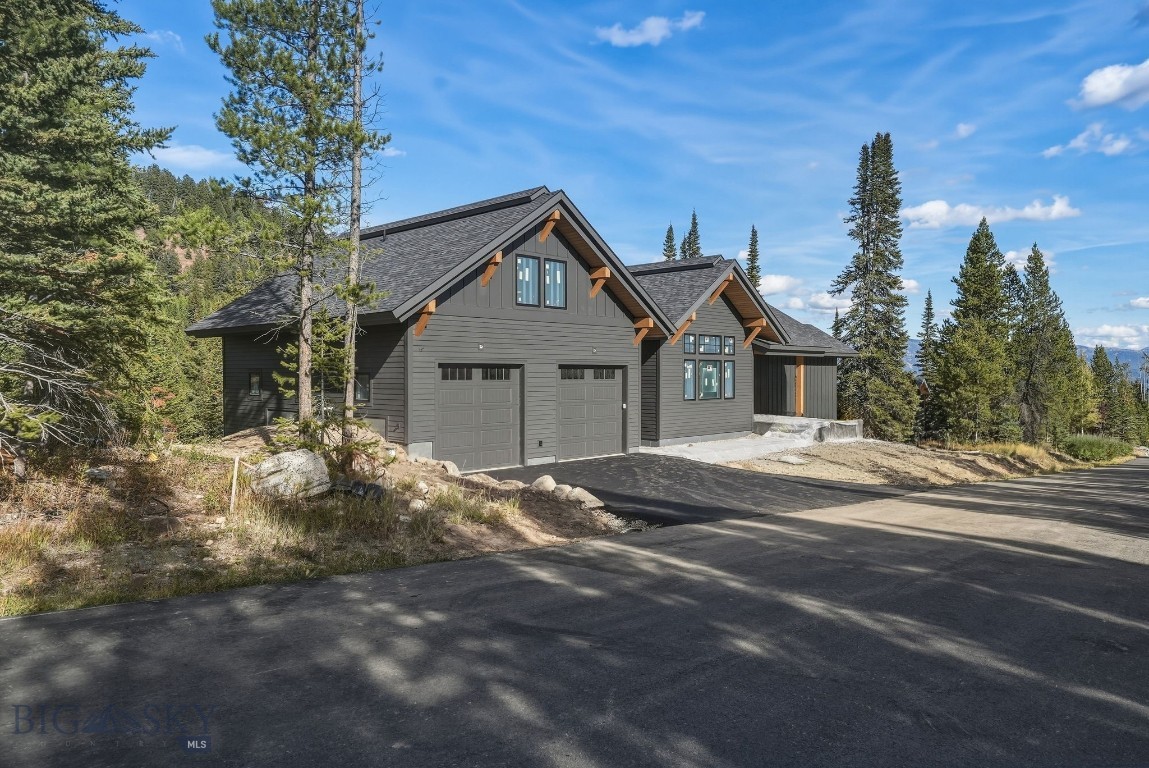 Dark gray mountain-style home with two garage doors, steep gabled roofs, and a paved driveway among evergreens.