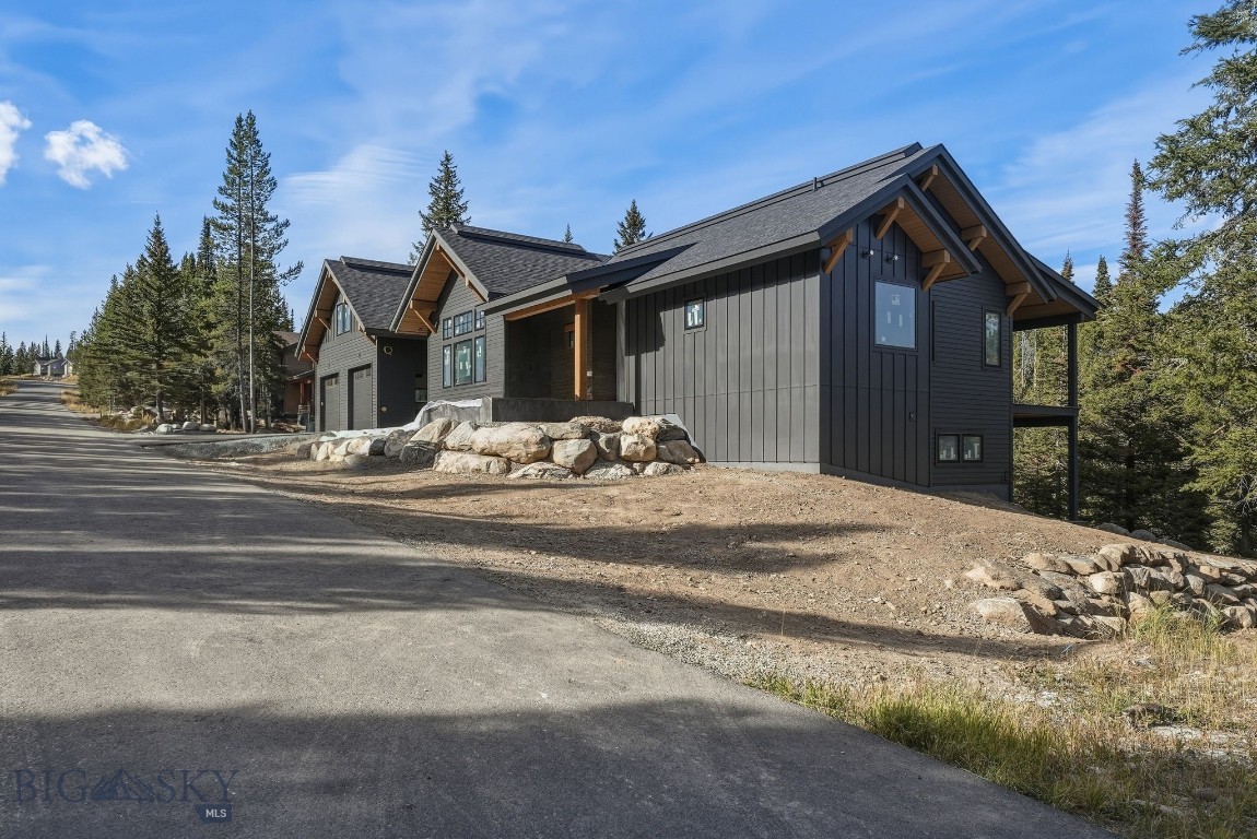 Dark gray mountain home with timber accents and a rock retaining wall beside a paved road.