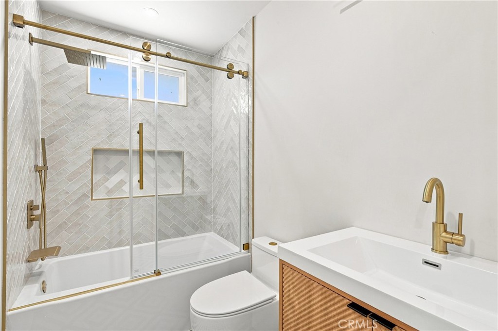 Bathroom with a glass-enclosed tub, pale herringbone tile, and wood-front vanity with gold fixtures.