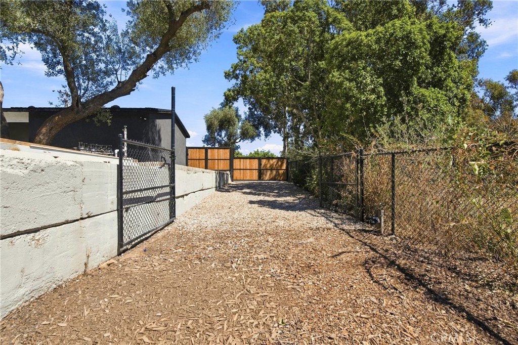 Fenced wood-chip access lane with chain-link fencing, a low wall, and a wooden gate.