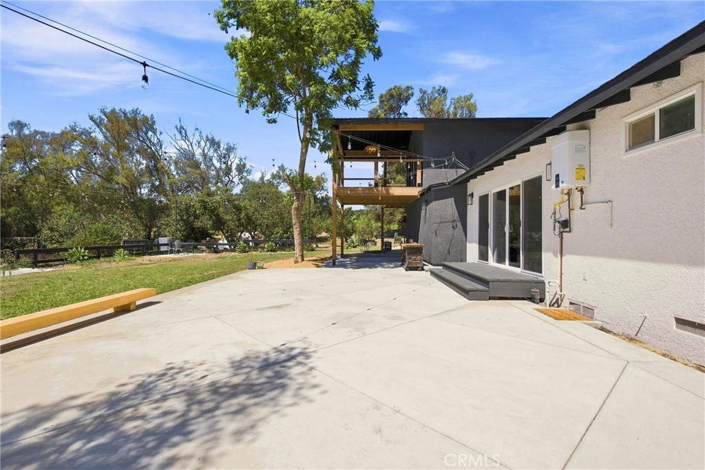 Concrete patio beside the home, with sliding doors and a raised wooden balcony overlooking a tree-lined lawn.