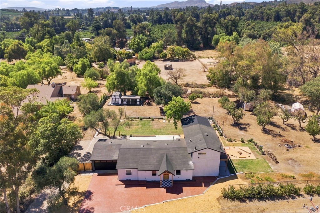 Aerial view of a home with a paved patio, detached outbuilding, fenced paddocks, and wooded hills.