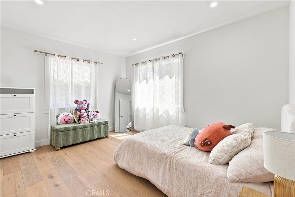 Bedroom with a low bed, plush toys on a green bench, and tall curtained windows.