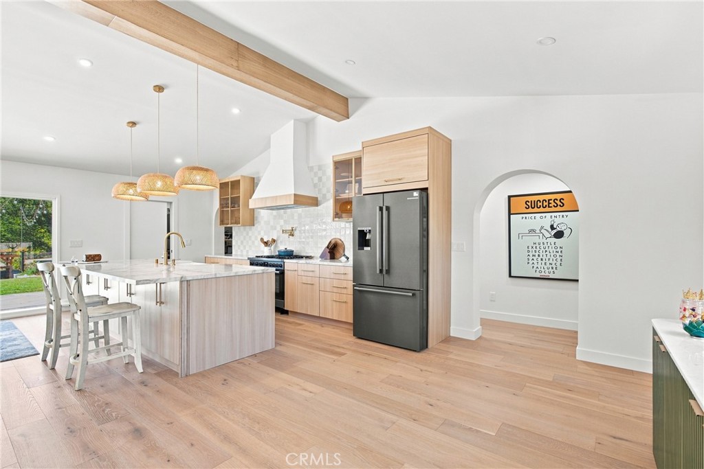 Modern kitchen with a large island, light wood cabinets, stainless steel refrigerator, and exposed ceiling beam.