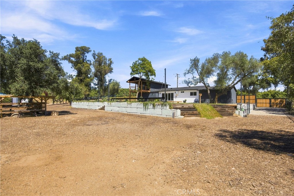 Single-story home behind a broad dirt yard, with a raised patio, mature trees, and wooden fencing.