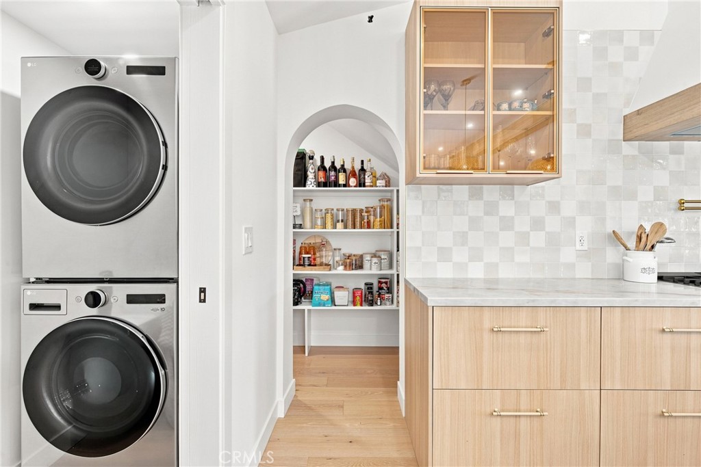 Kitchen with stacked washer and dryer beside a stocked arched pantry and light wood cabinetry.
