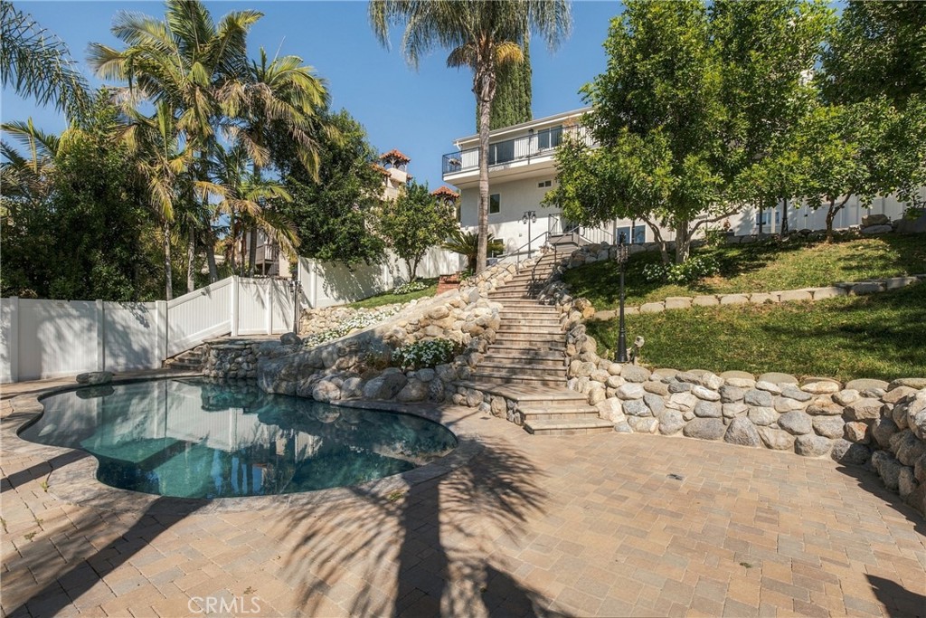 Backyard pool with stone steps, rock walls, palms, and a white two-story home.