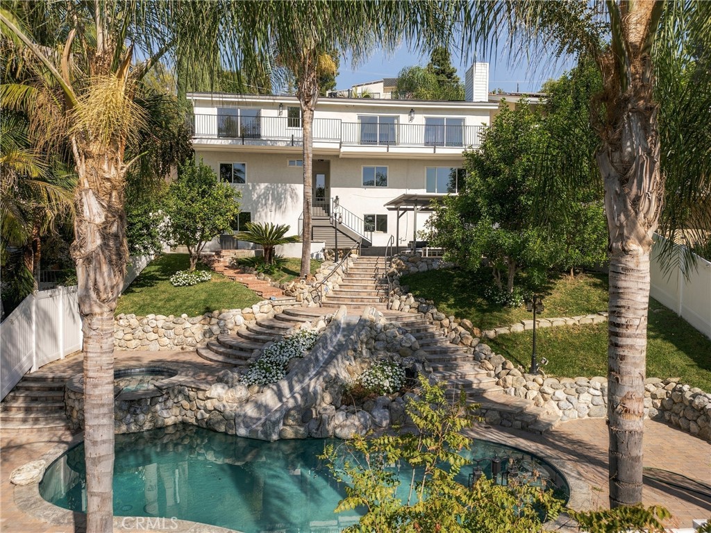 Two-story home behind a landscaped pool area, with stone steps, retaining walls, and palm trees.