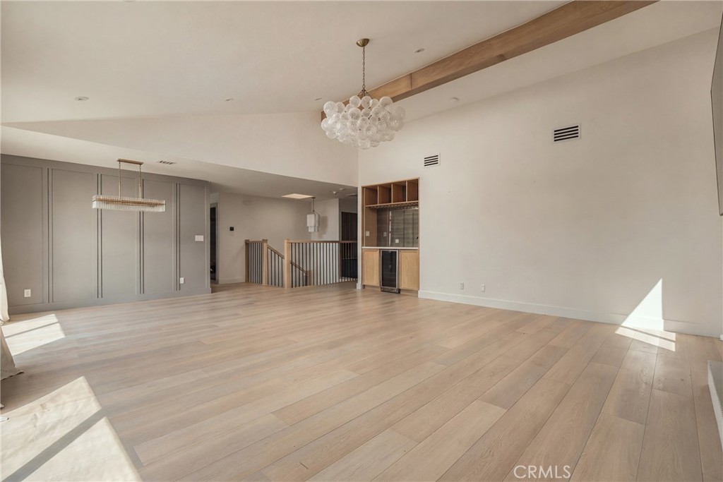 Vacant open living and dining area with pale wood floors, chandeliers, and a built-in bar.