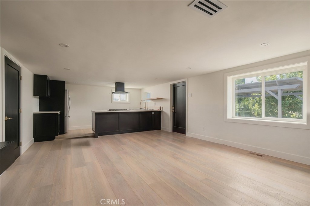 Vacant open-plan kitchen and living area with dark cabinetry, light wood floors, and a large window.