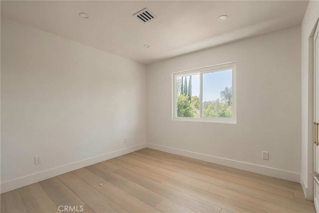 Vacant room with light wood flooring, recessed lights, and a window overlooking trees.
