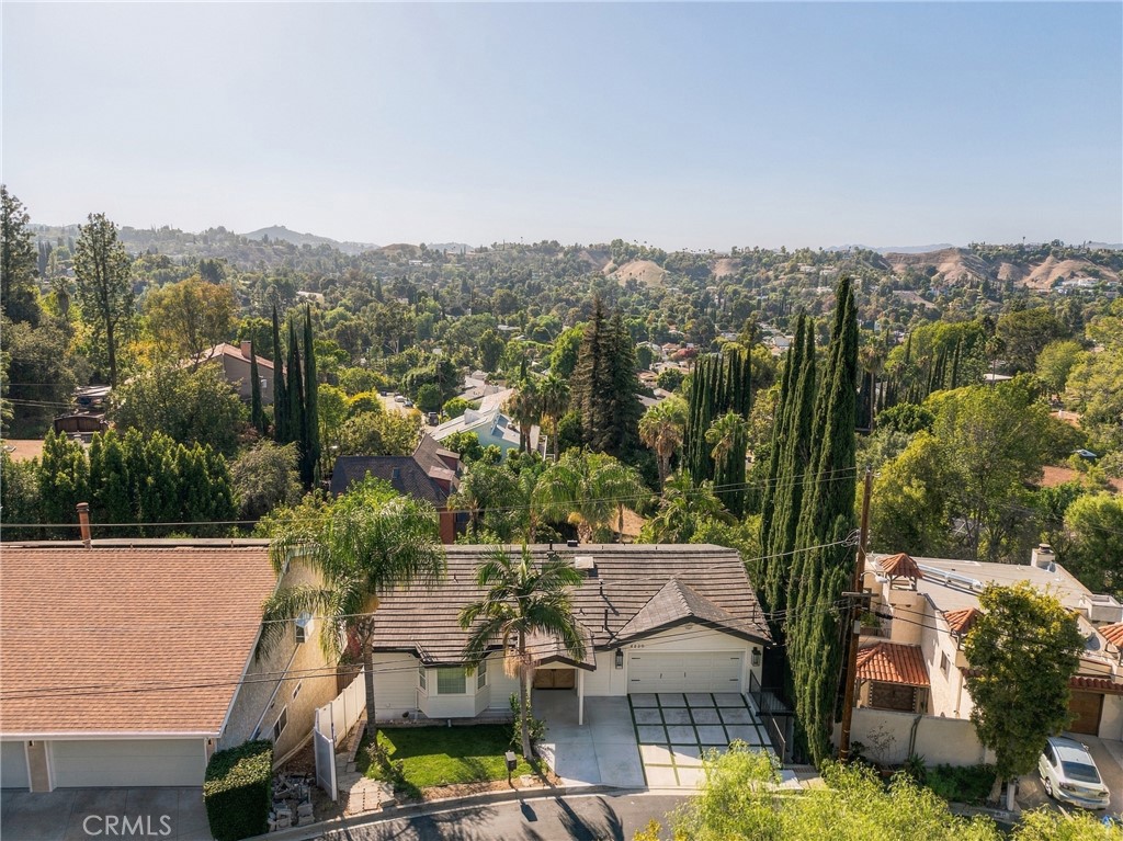Aerial view of a single-story home, paved driveway, palm trees, and wooded hills.