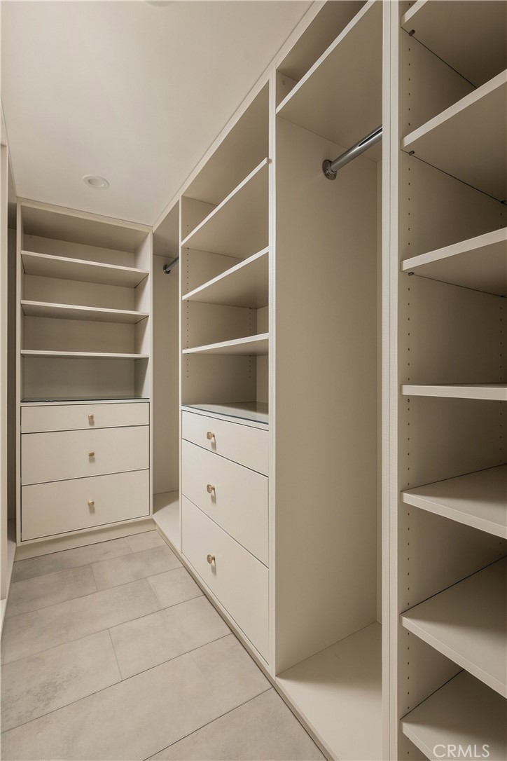 Walk-in closet with white built-in shelves, hanging rods, drawers, and large tiled flooring.