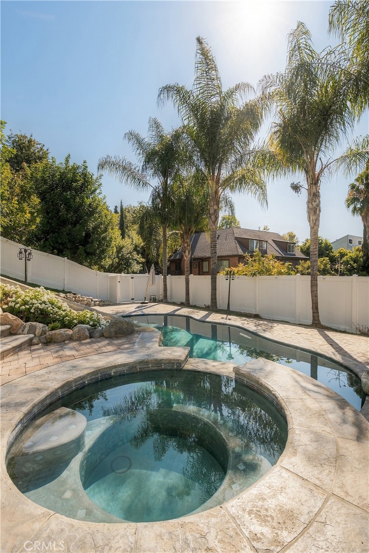 Backyard pool and circular spa with a stone patio, white fence, and tall palm trees.