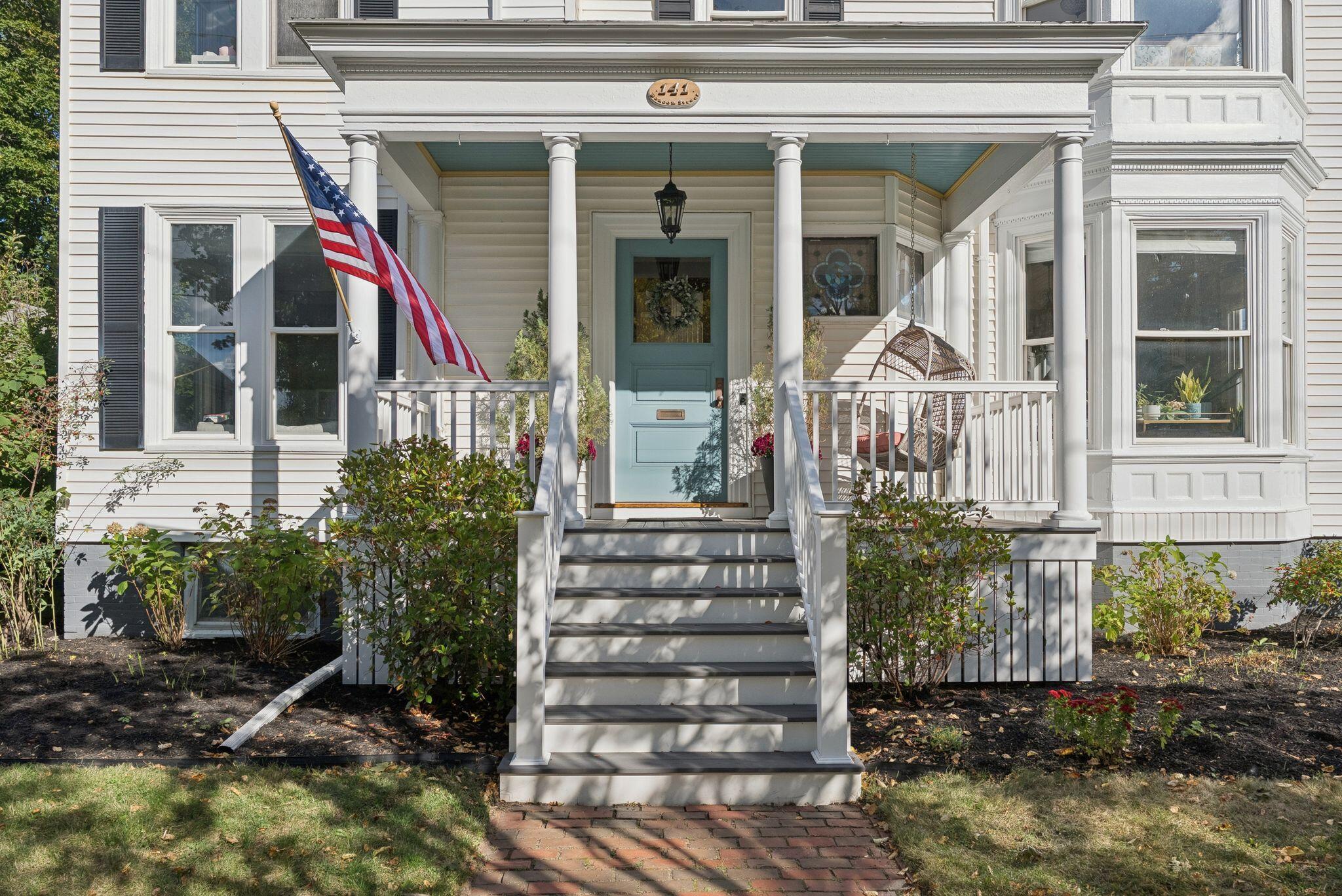 White house with a columned porch, light blue door, American flag, and hanging wicker chair.