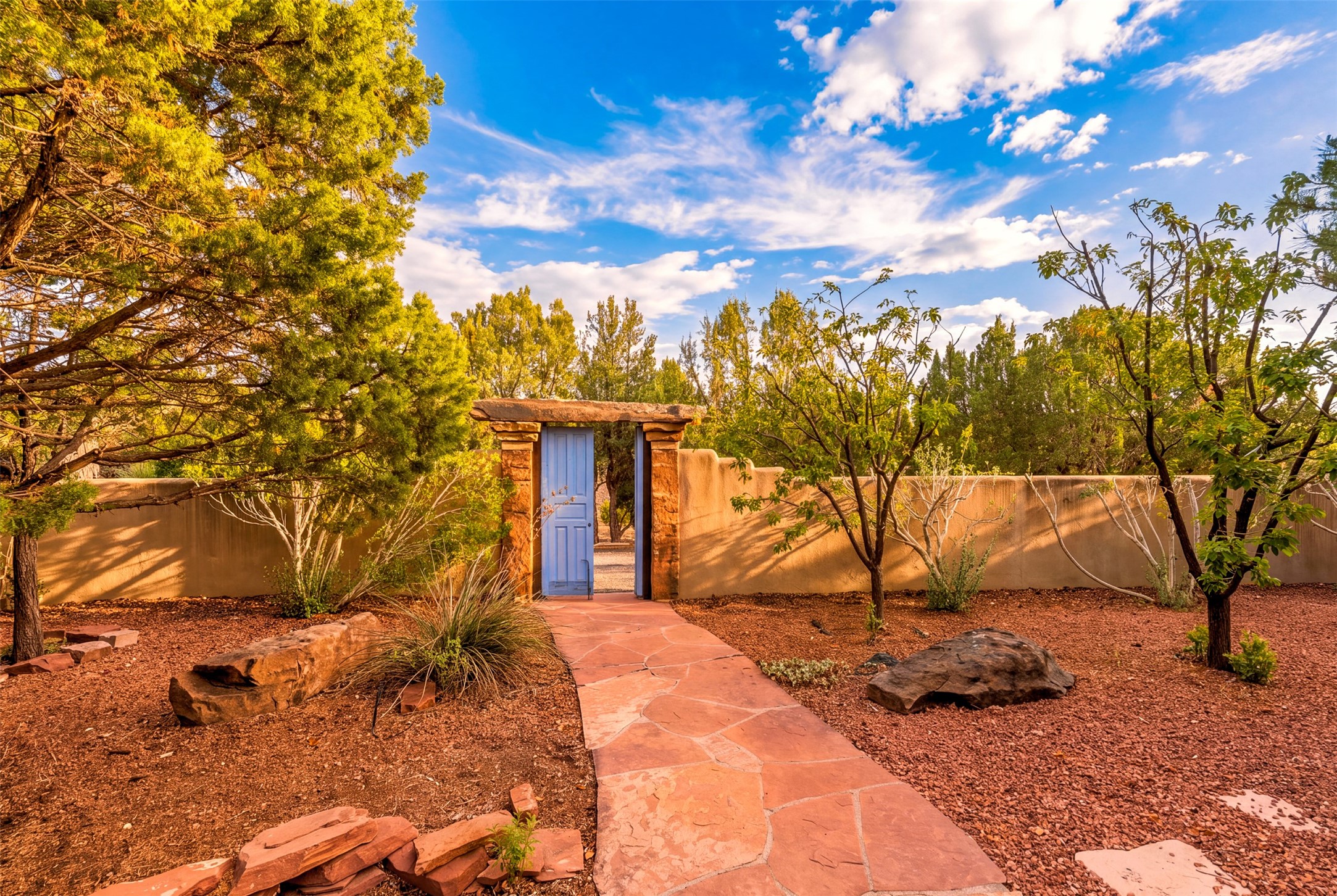 Stone walkway leading to a blue gate in an adobe garden wall, framed by mature trees and red gravel.