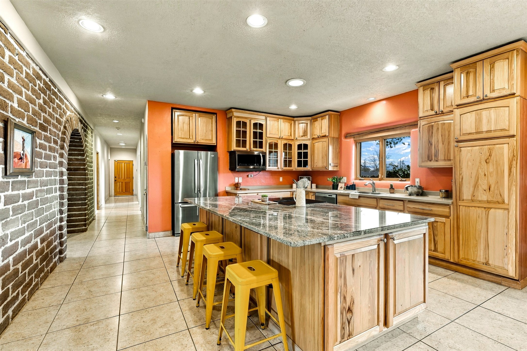 Kitchen with wood cabinets, a granite island, yellow stools, and a brick accent wall.