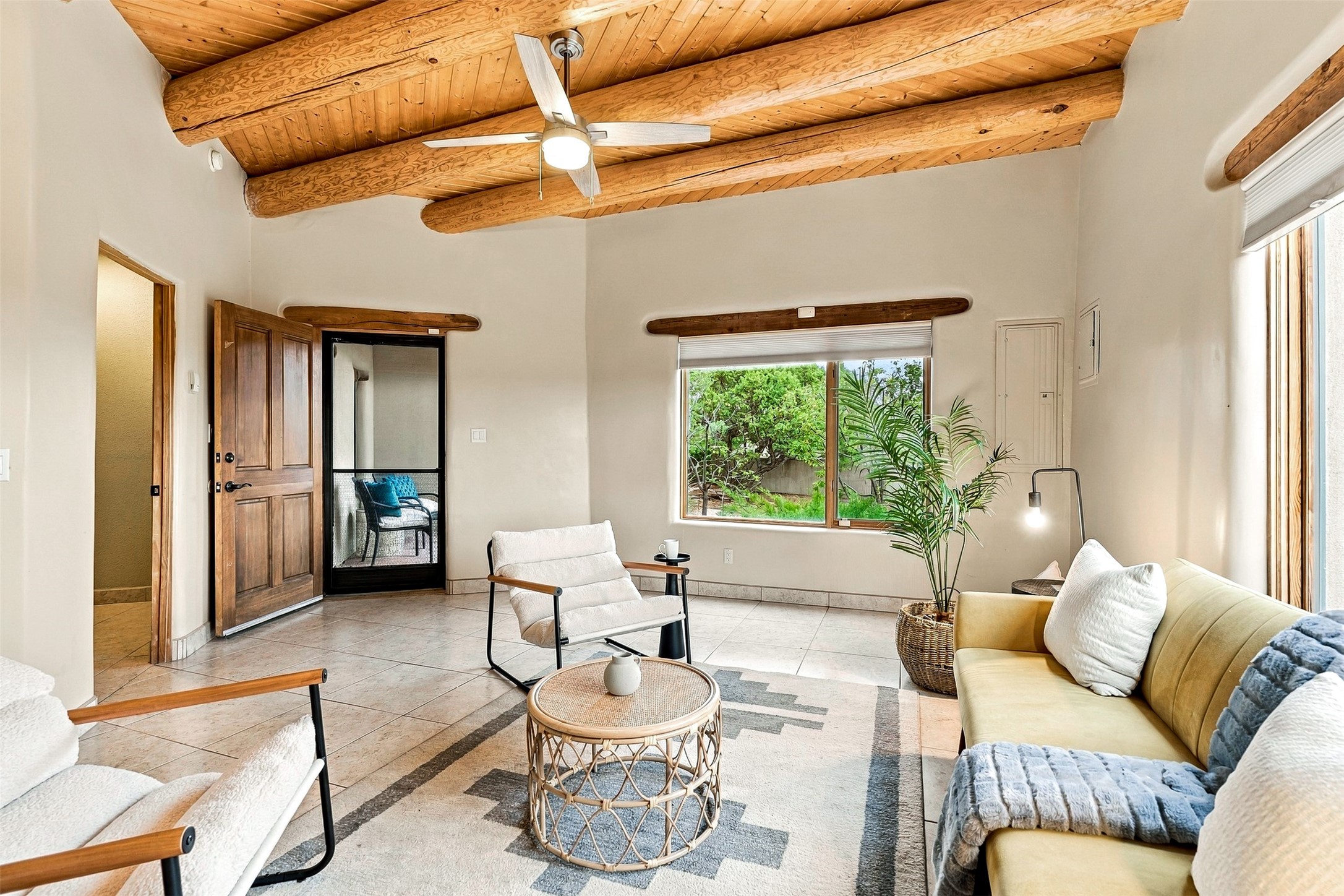 Living room with exposed wood ceiling beams, tiled floors, and a large window overlooking greenery.