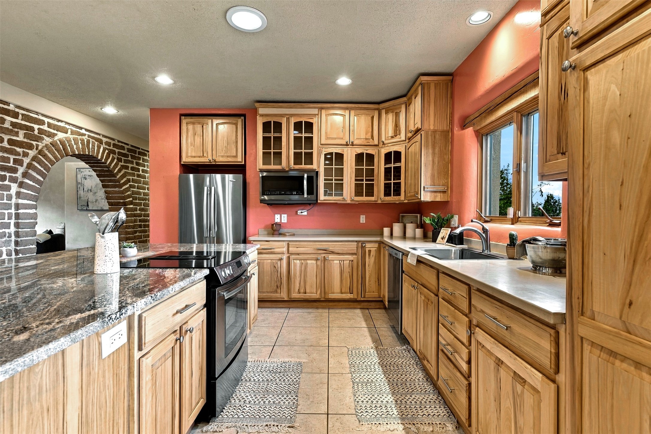 Kitchen with natural wood cabinets, granite island, stainless steel appliances, and a brick archway.