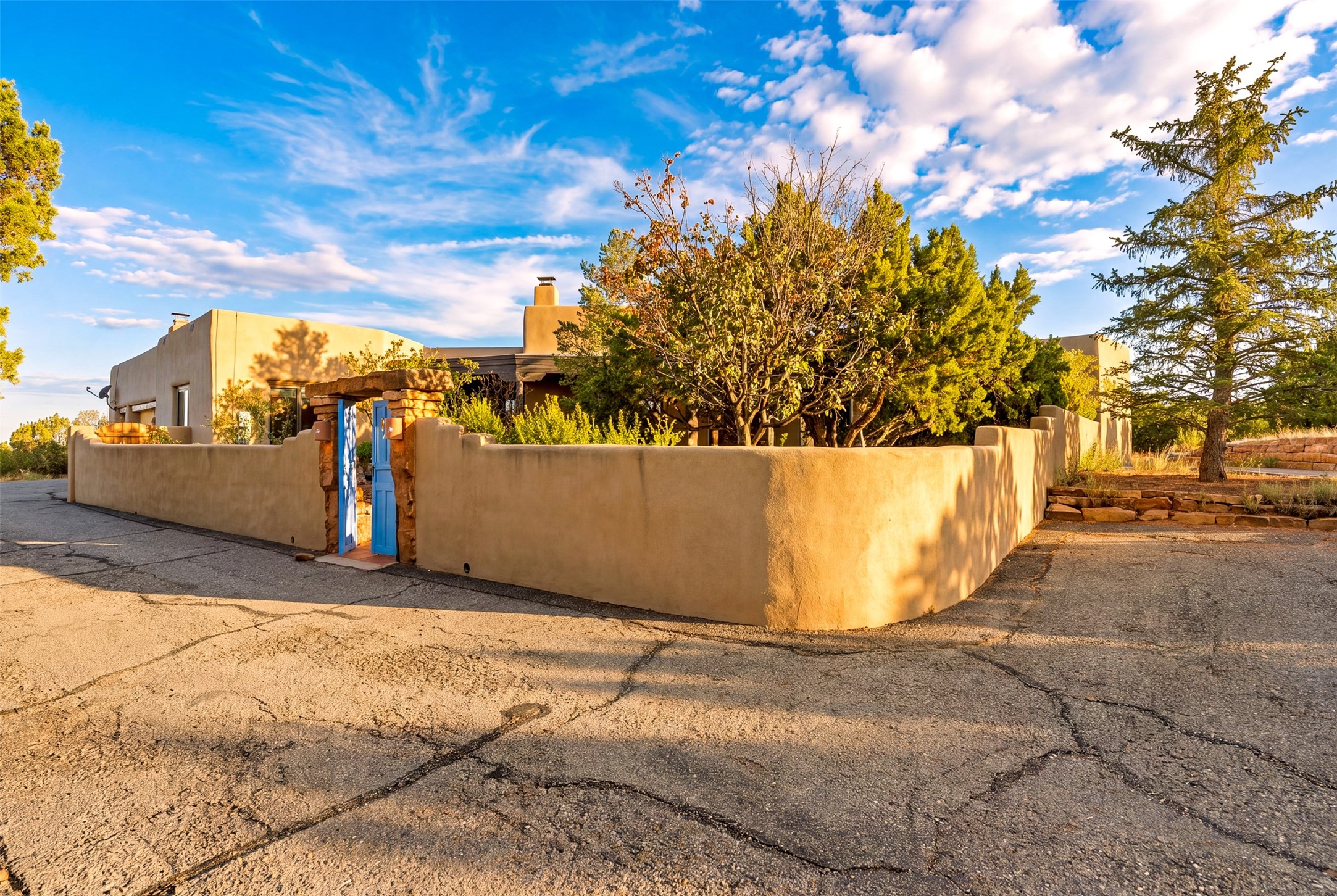 Santa Fe stucco home behind a tan wall, with a bright blue gate and mature trees.