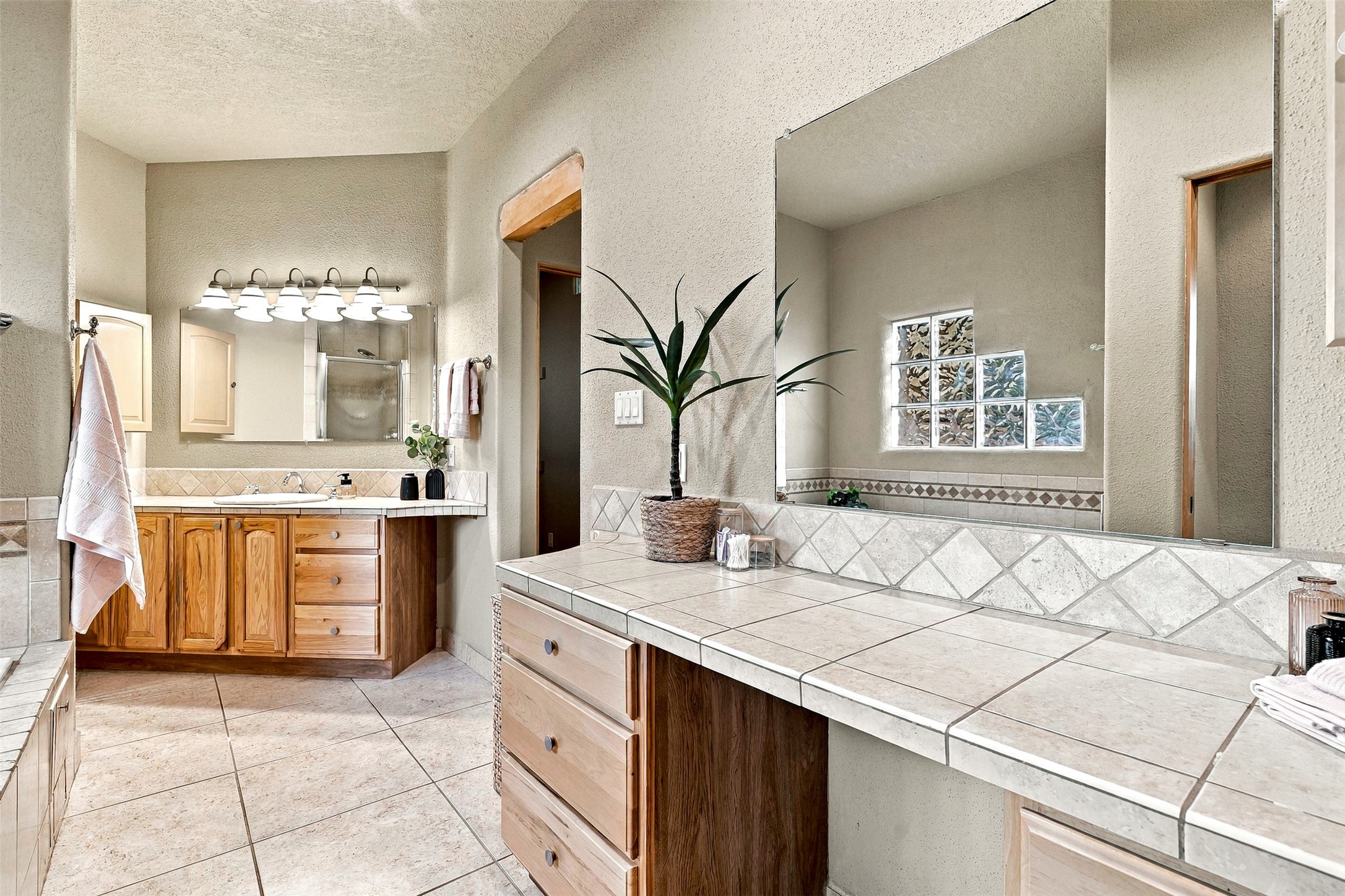 Bathroom with wood cabinetry, tiled counters, large mirrors, and a glass-block window.